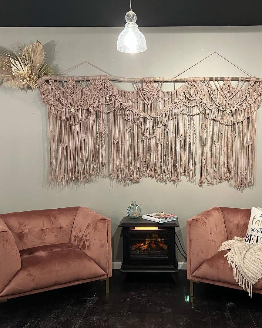 The waiting area: a large blush macrame wall hanging above two velvet chairs and a fireplace.