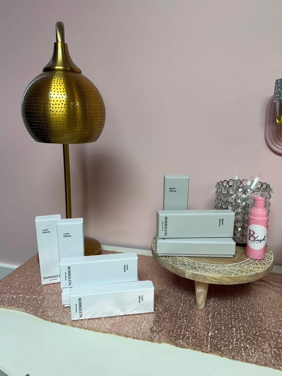 Lash and brow serums on a wooden riser beside a gold lamp.